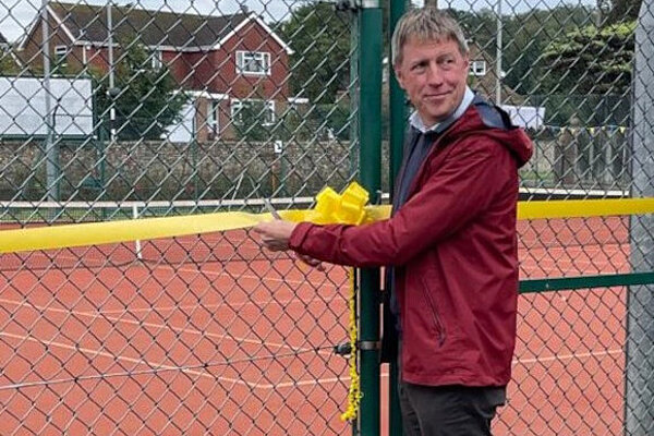 James is looking over his shoulder while holding scissors on a decoratively tied yellow ribbon across the gateway to a tennis court. He appears to be about to cut off his fingers.