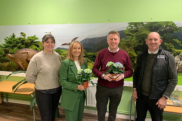 James posing with Sarah Osborne and two staff from Paradise Park, in front of a dinosaur tableau. James and Sarah are holding pos of flowers.