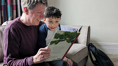 James on a sofa in a comfortable room, reading a book with a young child. Both James and the child look really excited at what they're reading.