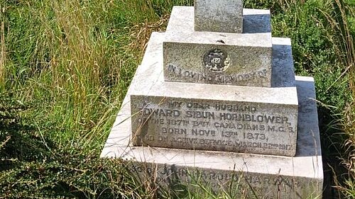 A headstone in the shape of a cross on a plinth, the stone weathered, and surrounded by untended grass and weeds. The inscription reads "In loving memory, my dear husband Edward Sibun Hornblower of the 87th Batt Canadians M.C.S. Born Novr 13th 1873 died on active service March 22nd 1917. Also of..." Further words are obscured by the grass.