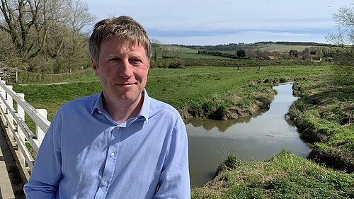 James MacCleary, in the foreground, on the South Downs Way near Alfriston, with a stream, fields, trees and blue sky behind him