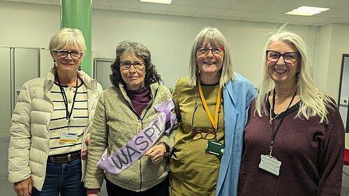 WASPI campaigners, one of whom is Councillor Chris Brett, posing for an indoor photo. One wears a sash with the word "WASPI" on it. They all look as if they mean business.