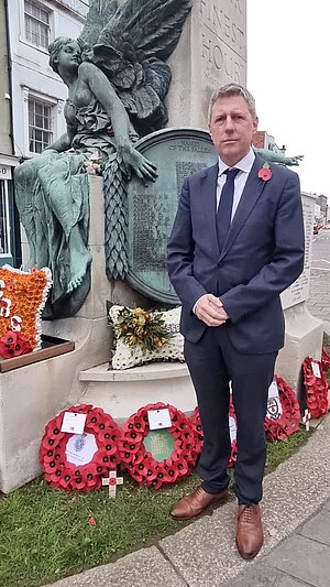 James in suit and dark tie by the Lewes war memorial, which has a number of wreathes at its base.