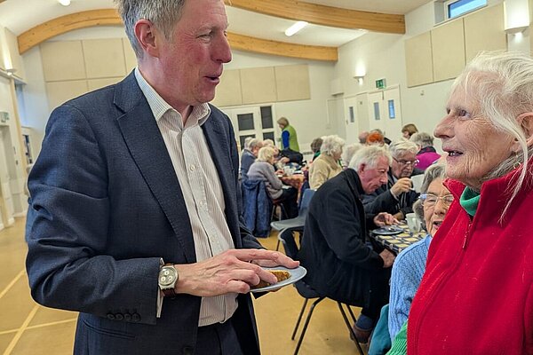 James in conversation in the foreground with a large hall behind. Other people are eating. James is snacking.