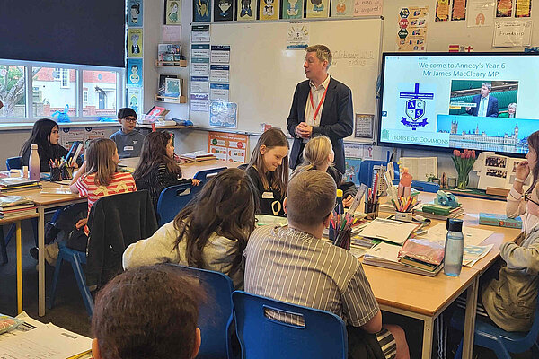 A busy classroom with a dozen children at tables and James at the front talking to them