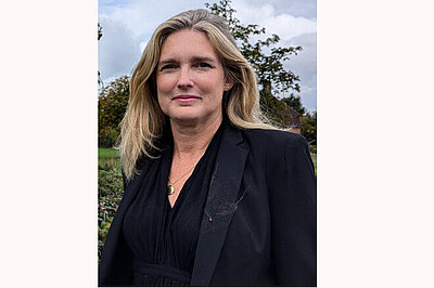 Portrait shot of Nadine Stothard, a white woman with long wavy blond hair. She is wearing a black v necked top and  black jacket with a gold pendant hanging from her neck.